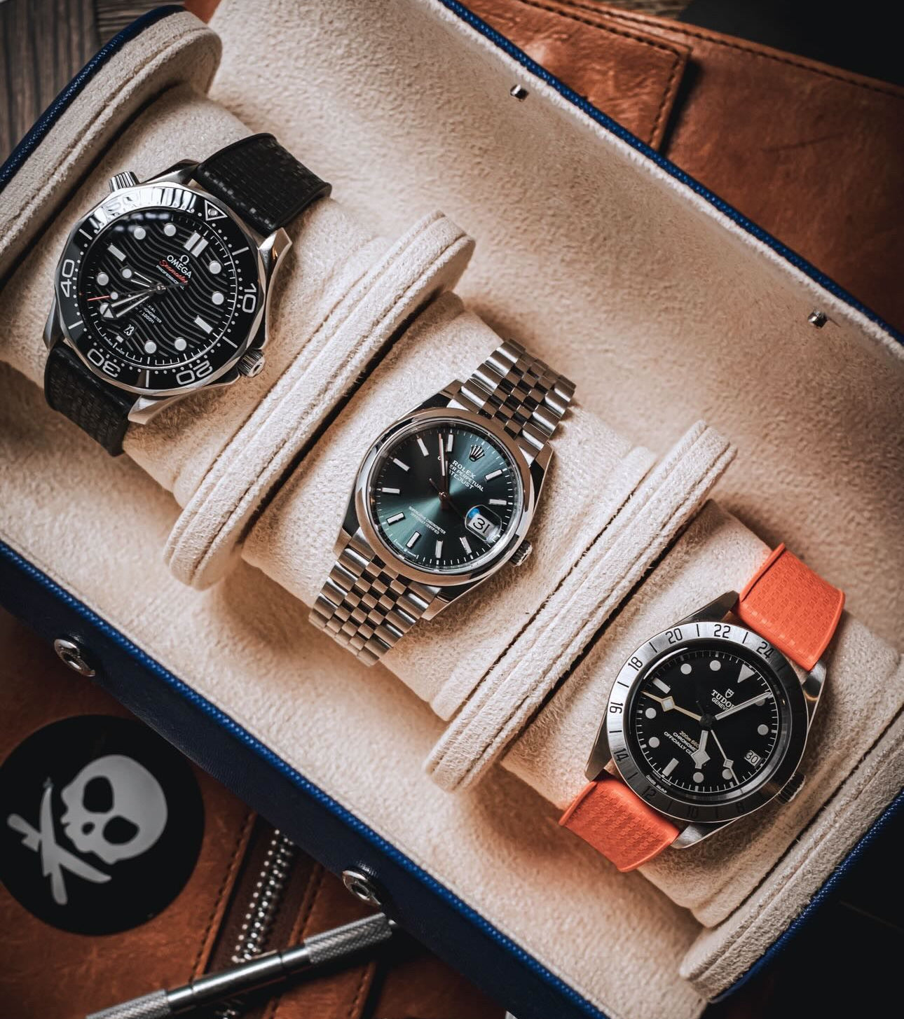 Three wristwatches displayed in a watch box with beige cushioning. The Omega on the left has a black dial and black WIS Watch Rubber Strap, the center watch is a Rolex Datejust that has a dark green dial and a silver bracelet, and the watch on the right is a Tudor has a black dial and an Orange Deployant Clasp WIS Watch Strap.