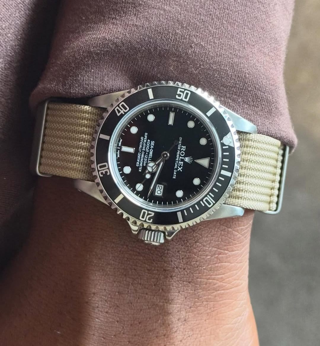 A Rolex Submariner watch with a black dial, black bezel, and a beige nylon WIS strap, shown worn on a wrist.