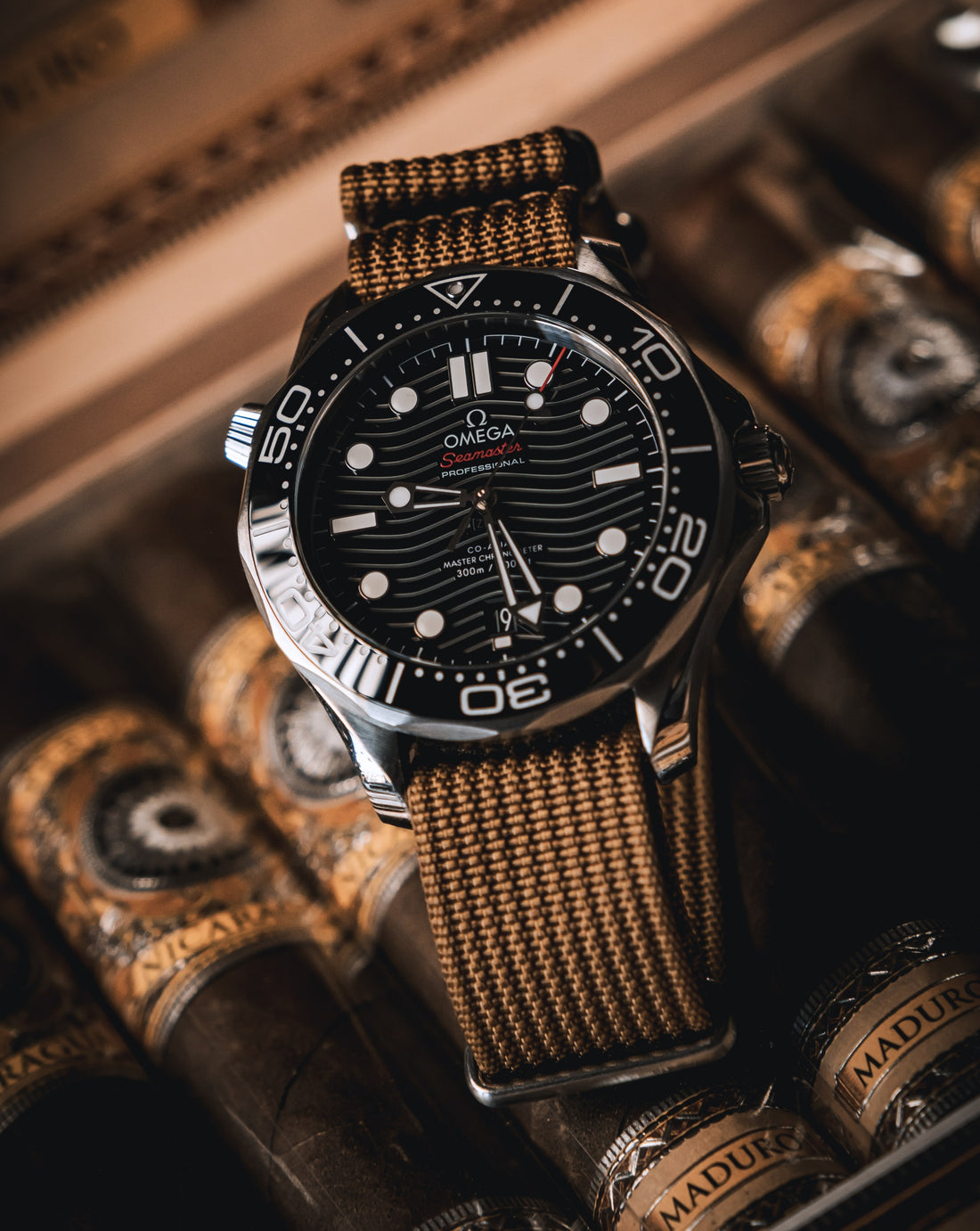 An Omega Seamaster wristwatch with a black dial, silver-toned case, and a brown woven nylon Nato WIS Watch strap, displayed resting on a row of cigars.