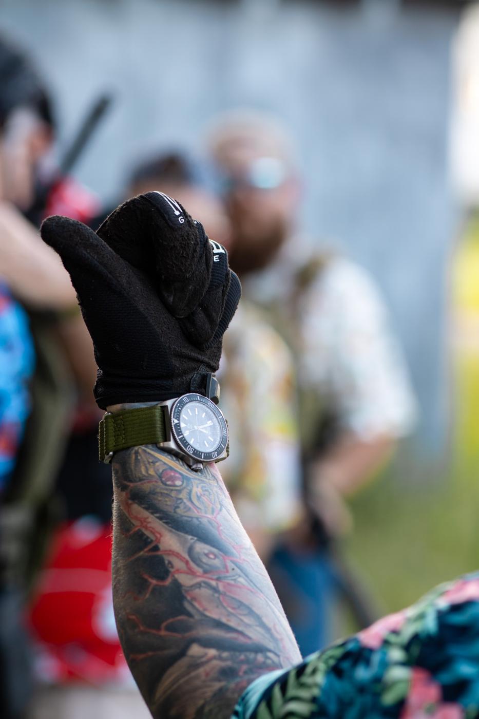 Tattooed arm wearing the SWCS on an olive Woven Nylon Velcro strap while holding a rifle